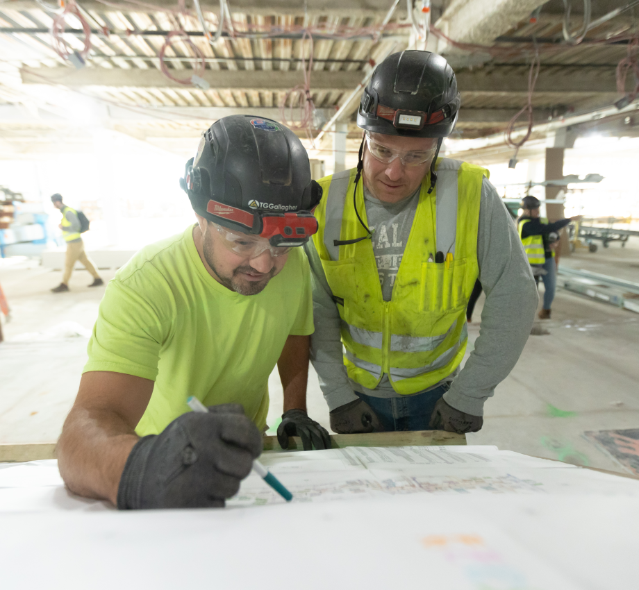 Two men writing at construction site, wearing hard hats and safety gear