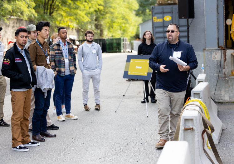 Man presenting to a group of people in front of a crane, outside